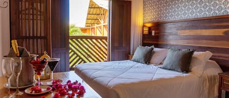 A hotel room with a large bed, a wooden headboard, a balcony with a view, and a table with a drink and flowers.