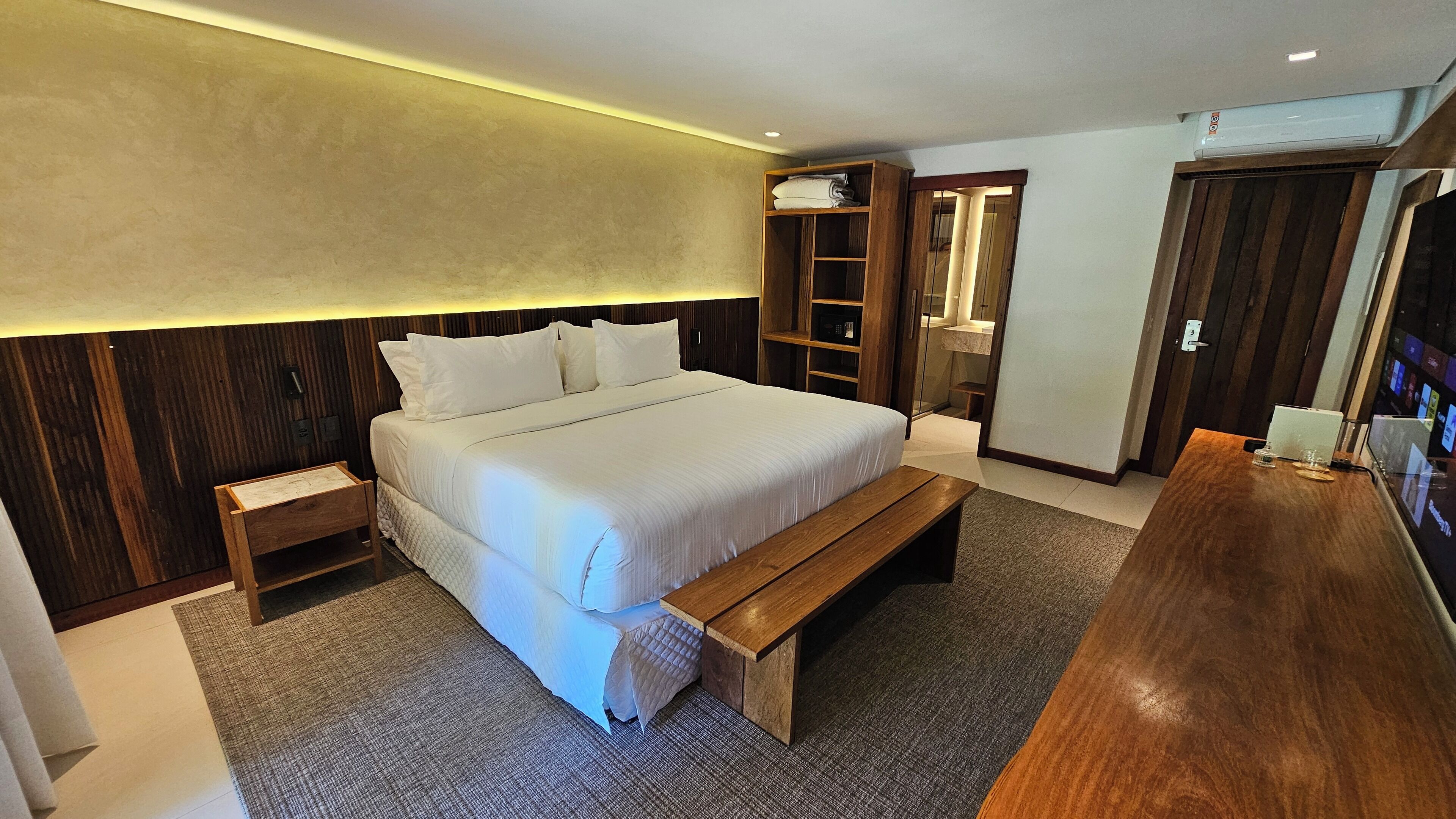 A modern hotel room with a large bed, wooden headboard, a wooden bench, a desk, and a wardrobe.