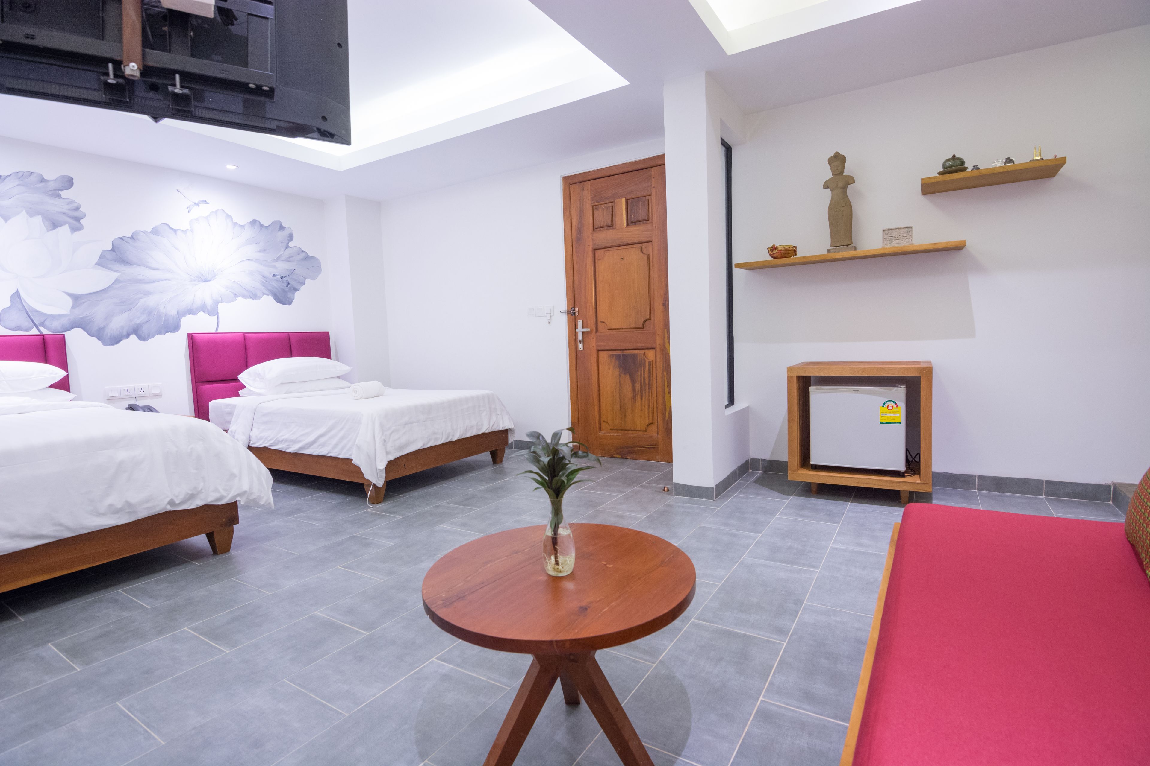 A hotel room with two beds, a small table with a vase, a wooden cabinet, and a wall-mounted shelf with decorative items.