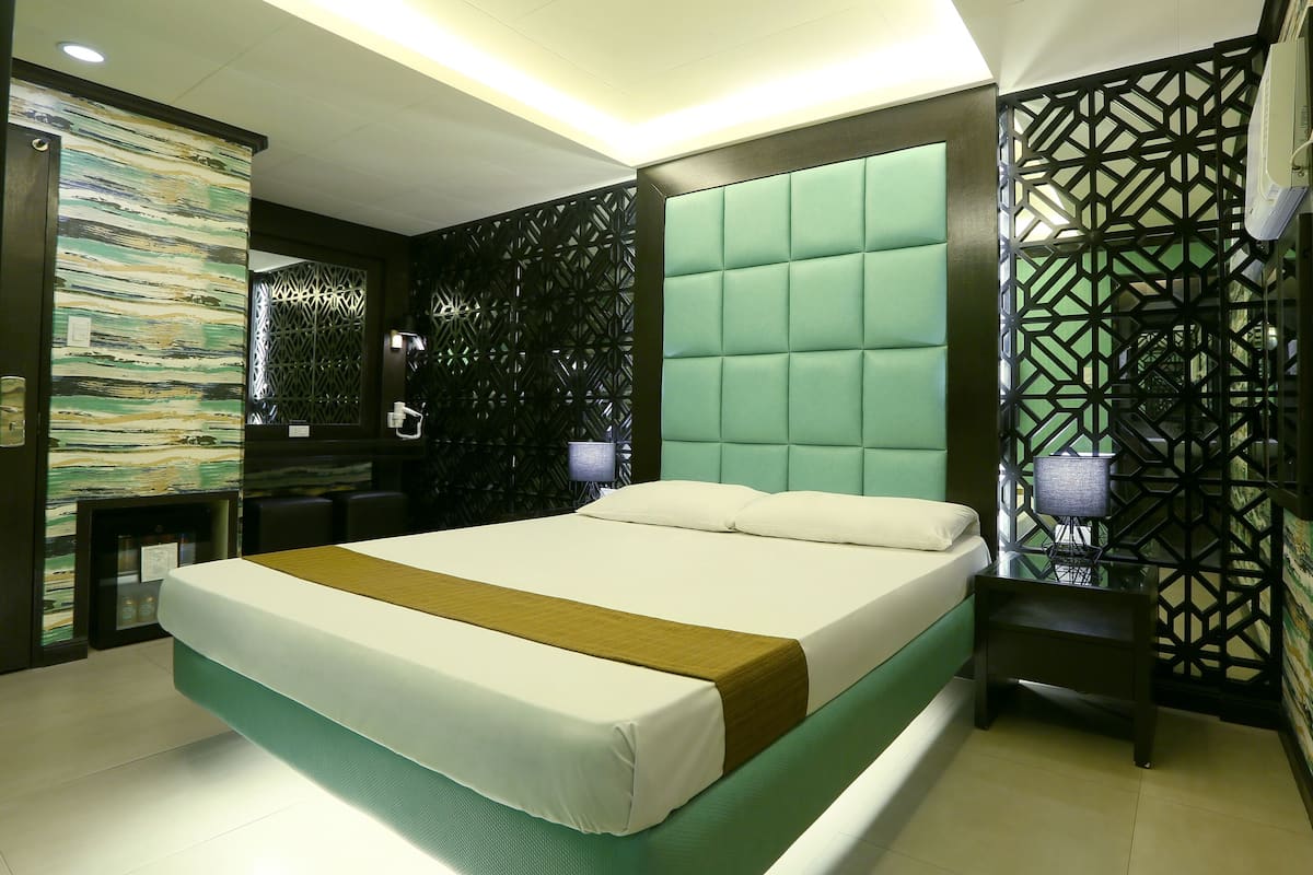 A modern hotel room with a large bed, a green upholstered headboard, and a geometric patterned wall.