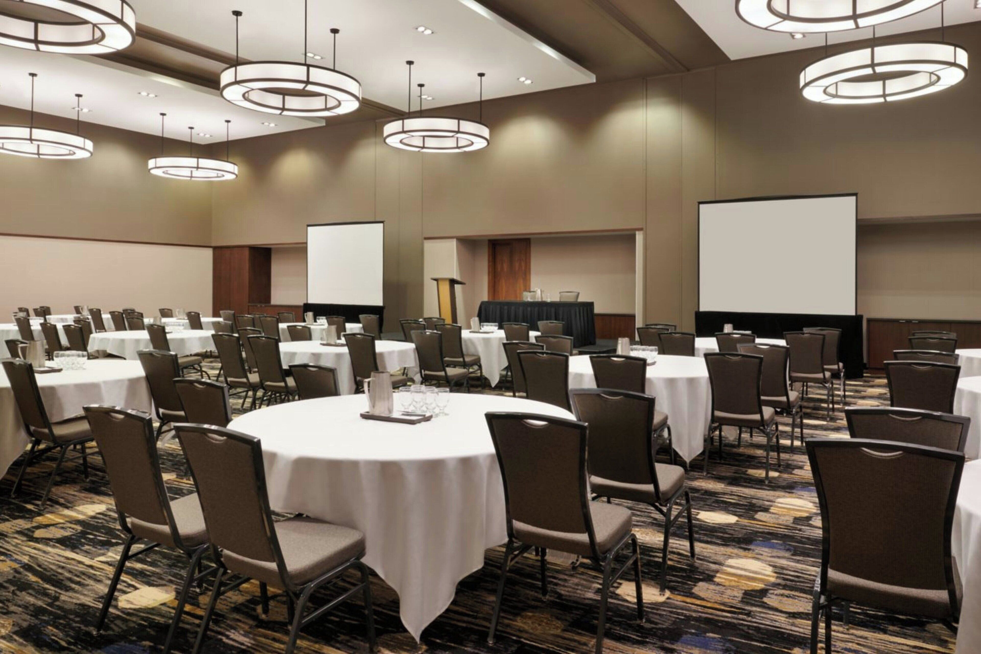 Un salón de conferencias con mesas redondas, sillas y una pantalla para proyector.