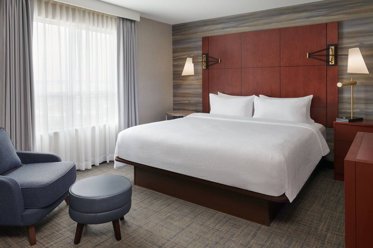 Une chambre d’hôtel avec un grand lit, une chaise, un petit pouf et une table de chevet avec une lampe.