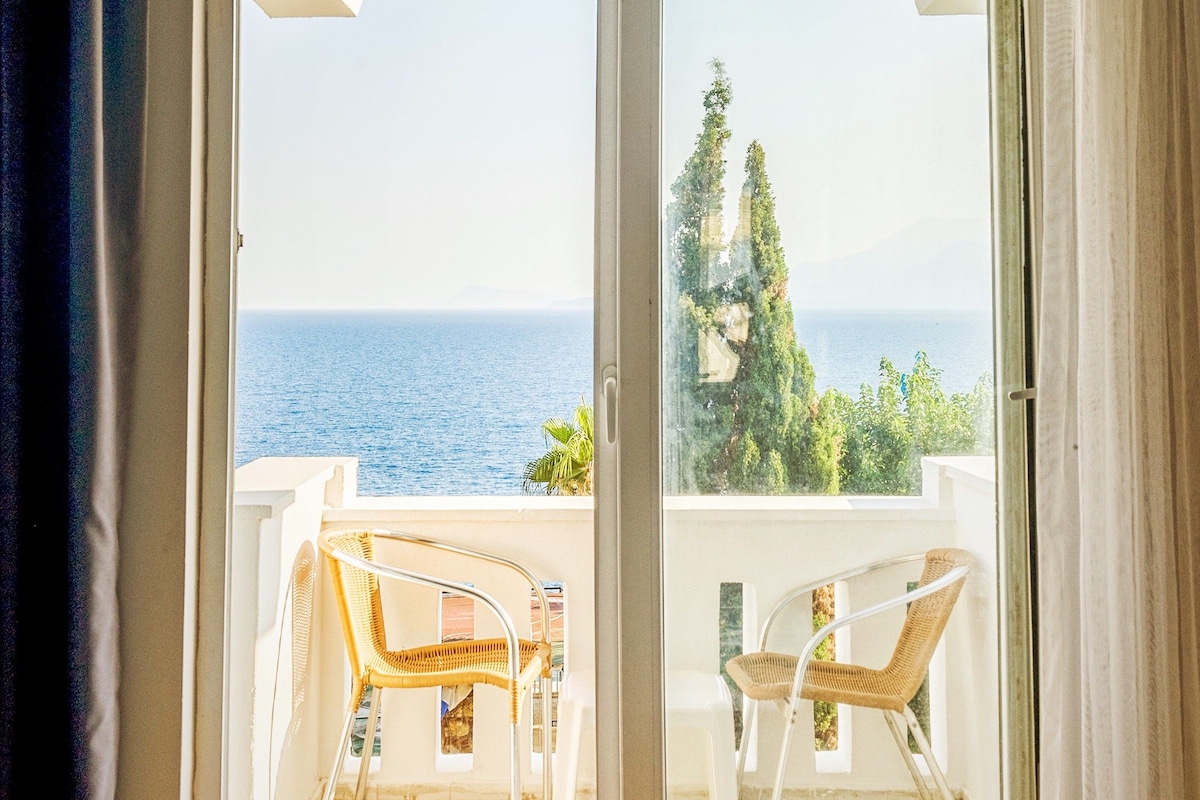 A room with a view of the sea, two wicker chairs, a bed, and a ceiling fan.