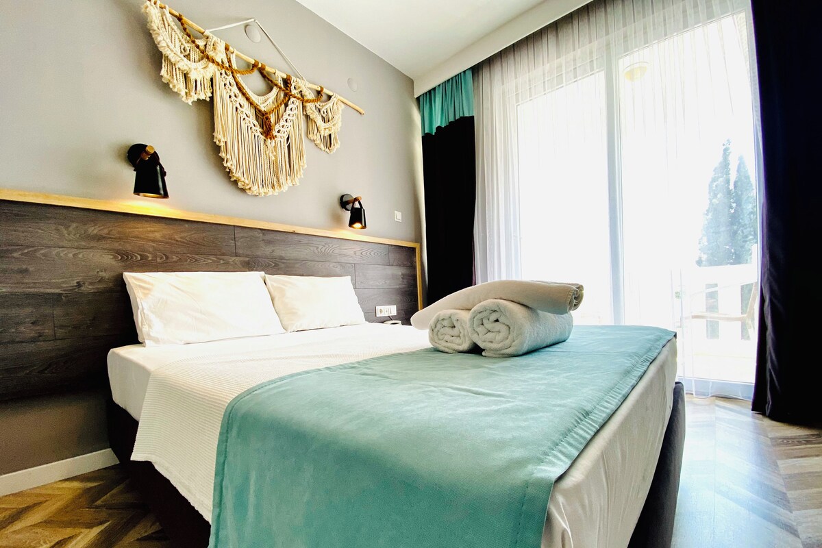 A hotel room with a bed, a macrame wall hanging, and a view of the outdoors.
