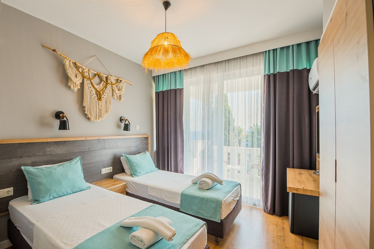 A hotel room with two beds, a wooden headboard, a hanging macrame wall decoration, a ceiling light, and a view of the outdoors through sheer curtains.