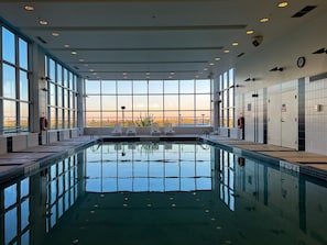 Una amplia piscina interior con agua cristalina y azul, rodeada de grandes ventanales con vistas a la ciudad.