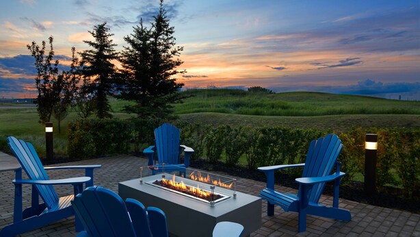 An outdoor seating area with Adirondack chairs and a fire feature, set against a backdrop of a sunset over a grassy field. - Courtyard by Marriott Calgary Airport (Calgary)