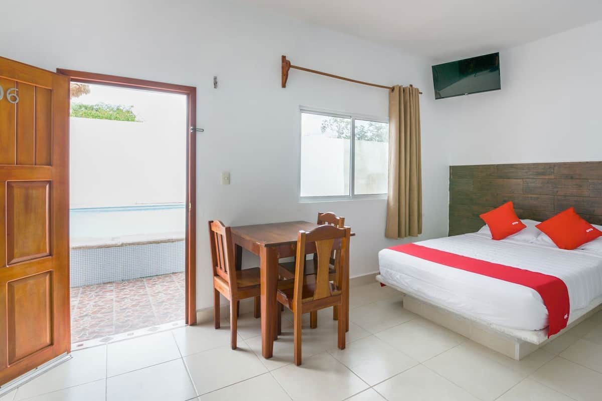 A room with a bed, a table with chairs, a TV mounted on the wall, and a window with curtains.