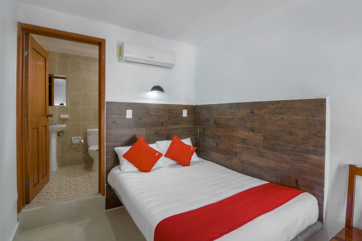 A hotel room with a wooden headboard, a bed with white bedding and red accents, a bathroom with a toilet and sink, and an air conditioning unit on the wall.