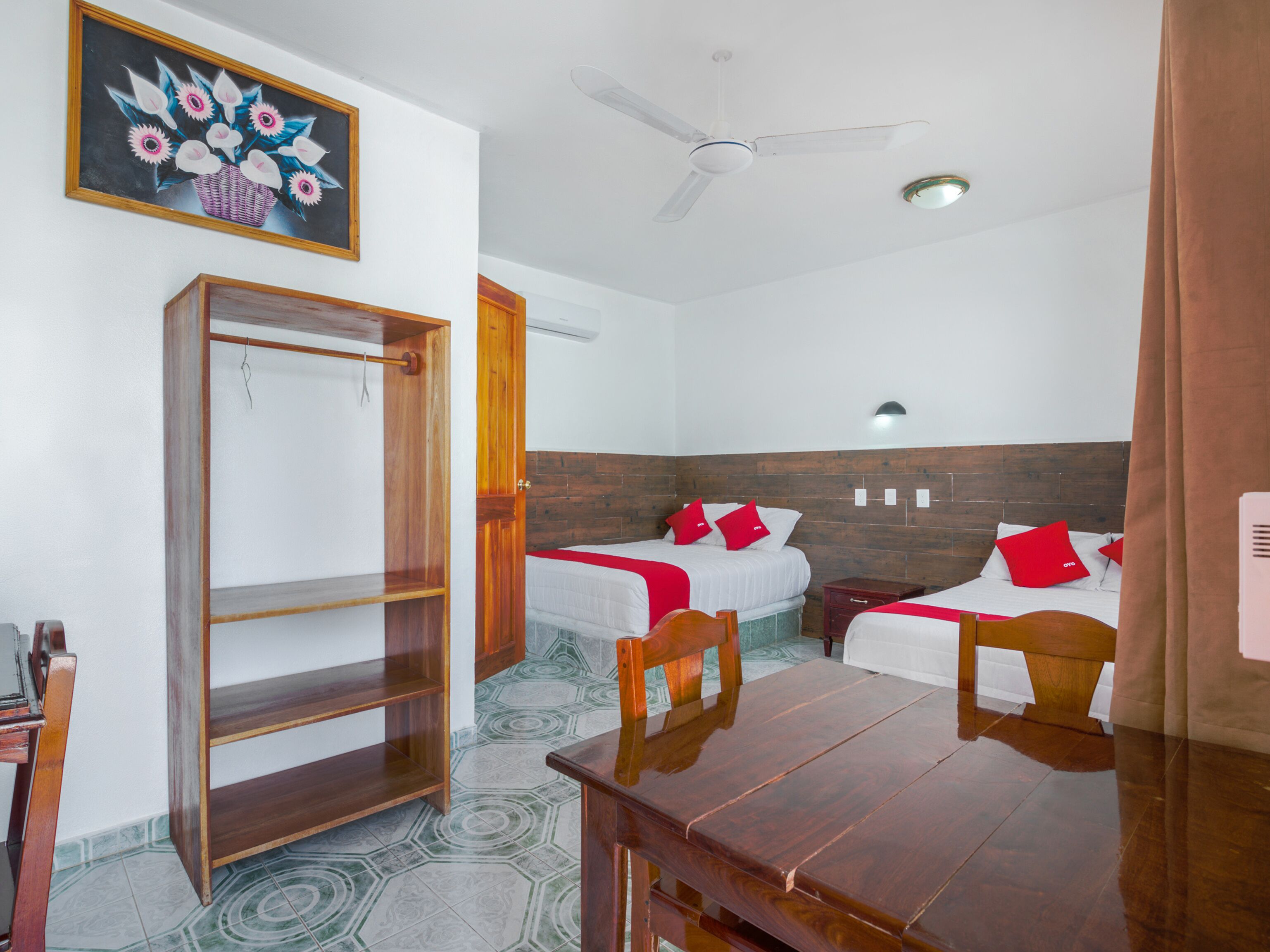 A hotel room with two beds, a wooden wardrobe, a dining table, and a ceiling fan.