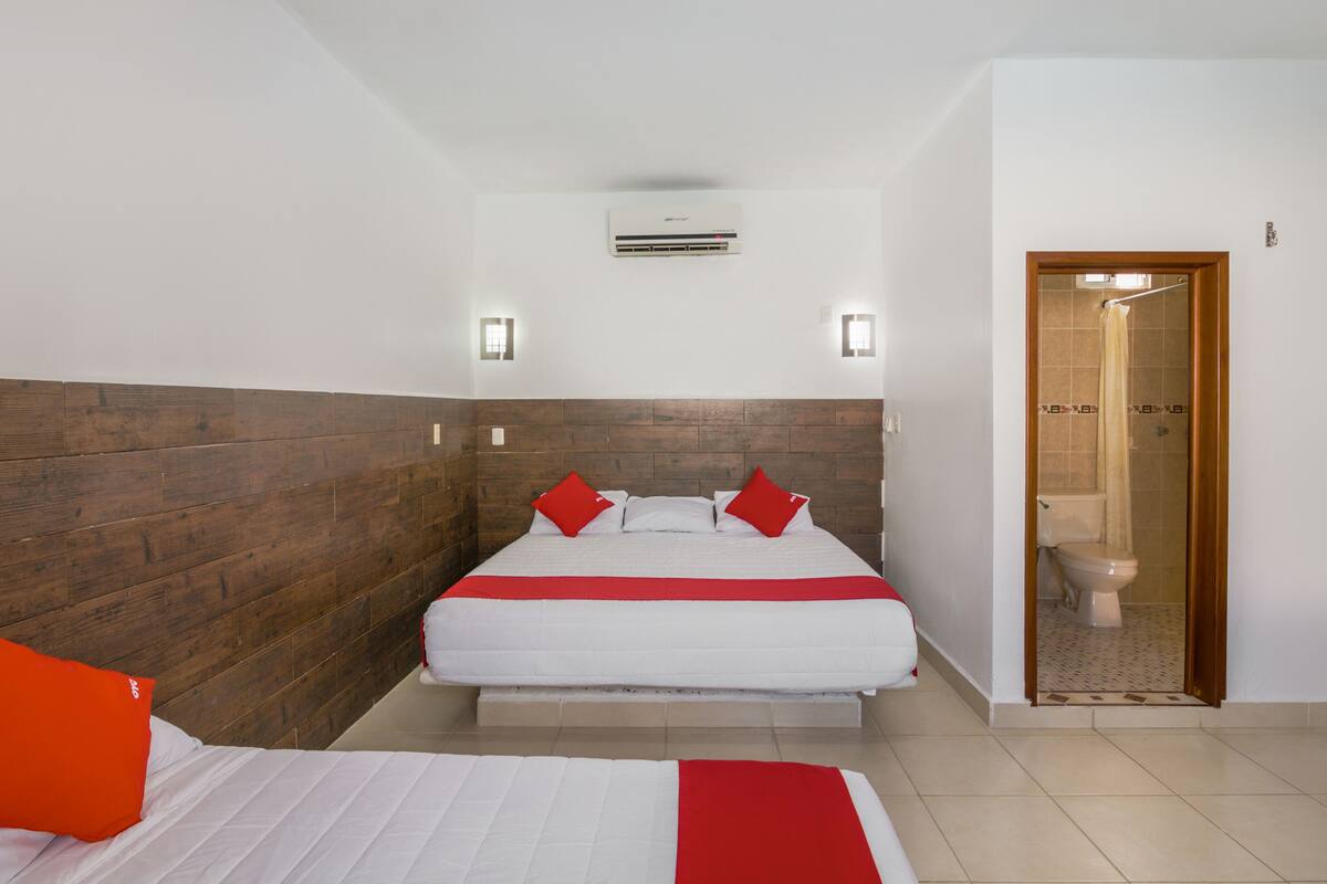 A hotel room with a wooden headboard, a bed with white bedding and red pillows, a small bedside table, and a shower area visible through an open door.