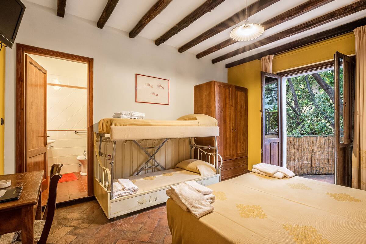 A dormitory room with a bunk bed, a single bed, a wooden wardrobe, a desk with a chair, and a window with a view of greenery.