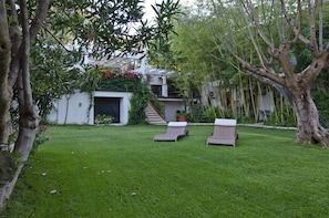 A lush garden with a white building, two lounge chairs, and a tree. - Hotel Santa Maria (Baunei)