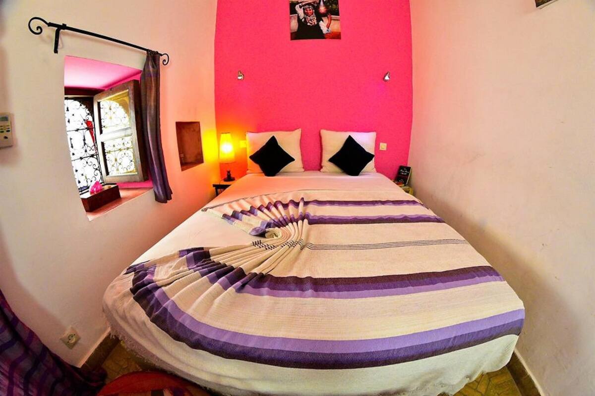 A bedroom with a bed, a window with curtains, a wall-mounted light, and a picture on the wall.