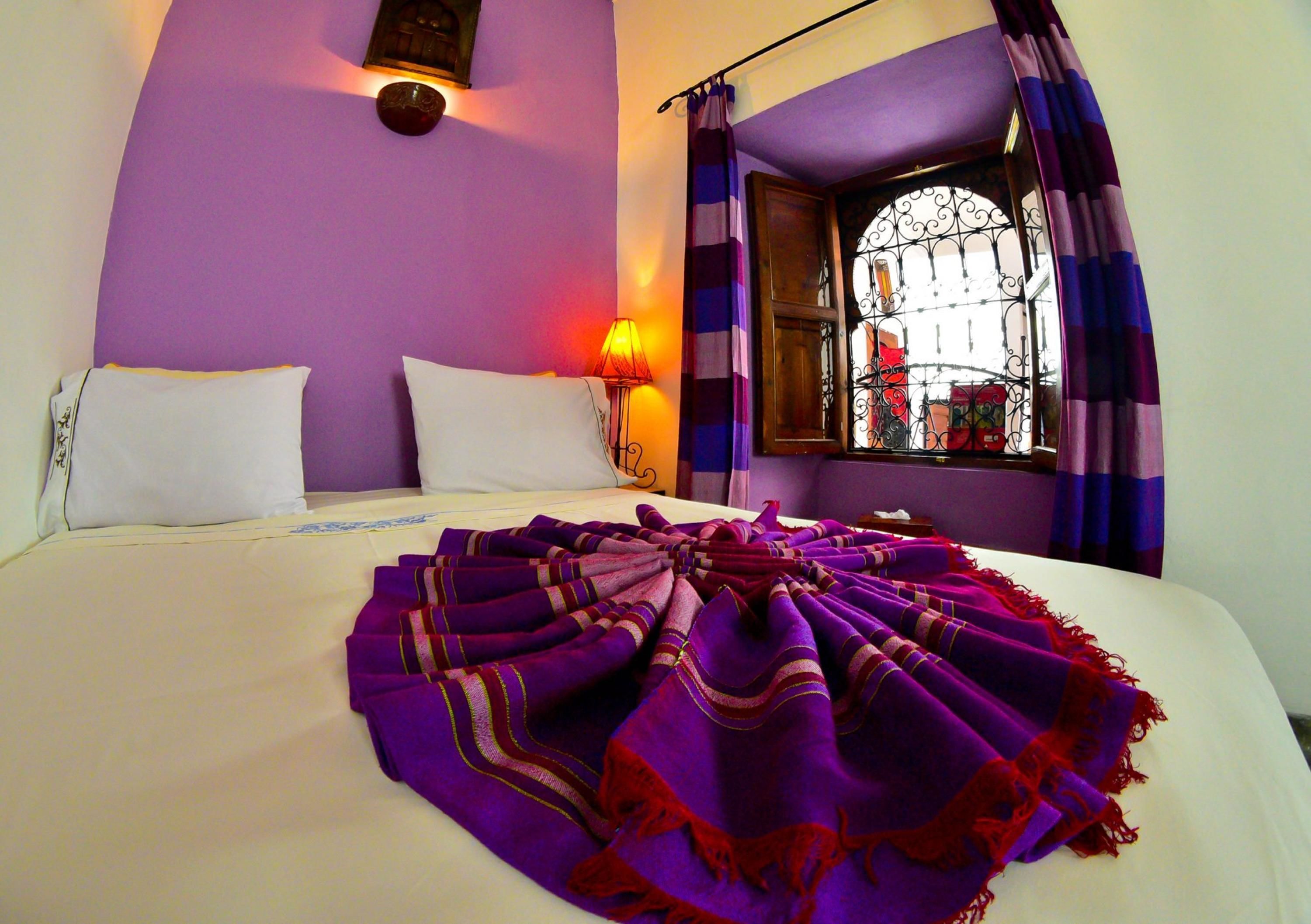 A bed with a purple blanket, white pillows, and a wall-mounted lamp.