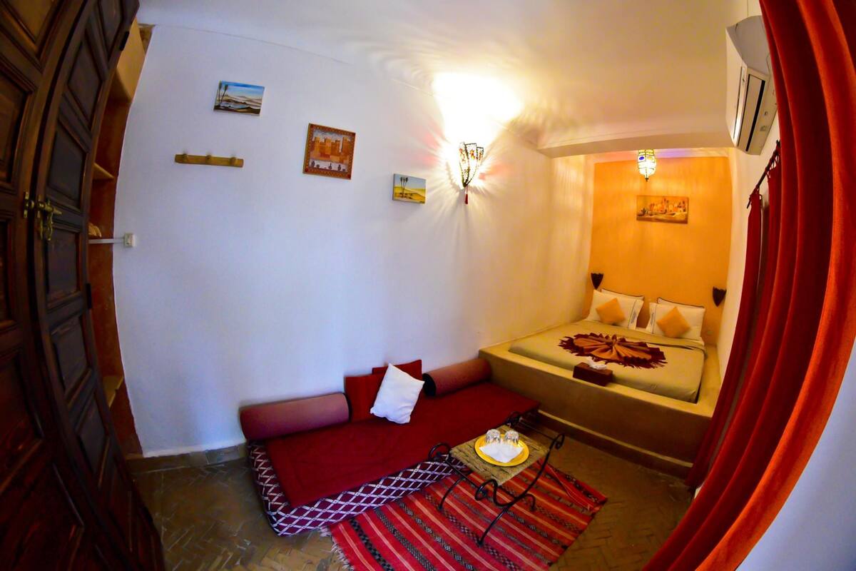 A compact bedroom with a bed, a small table with a tray, a red patterned rug, and a wall-mounted light fixture.