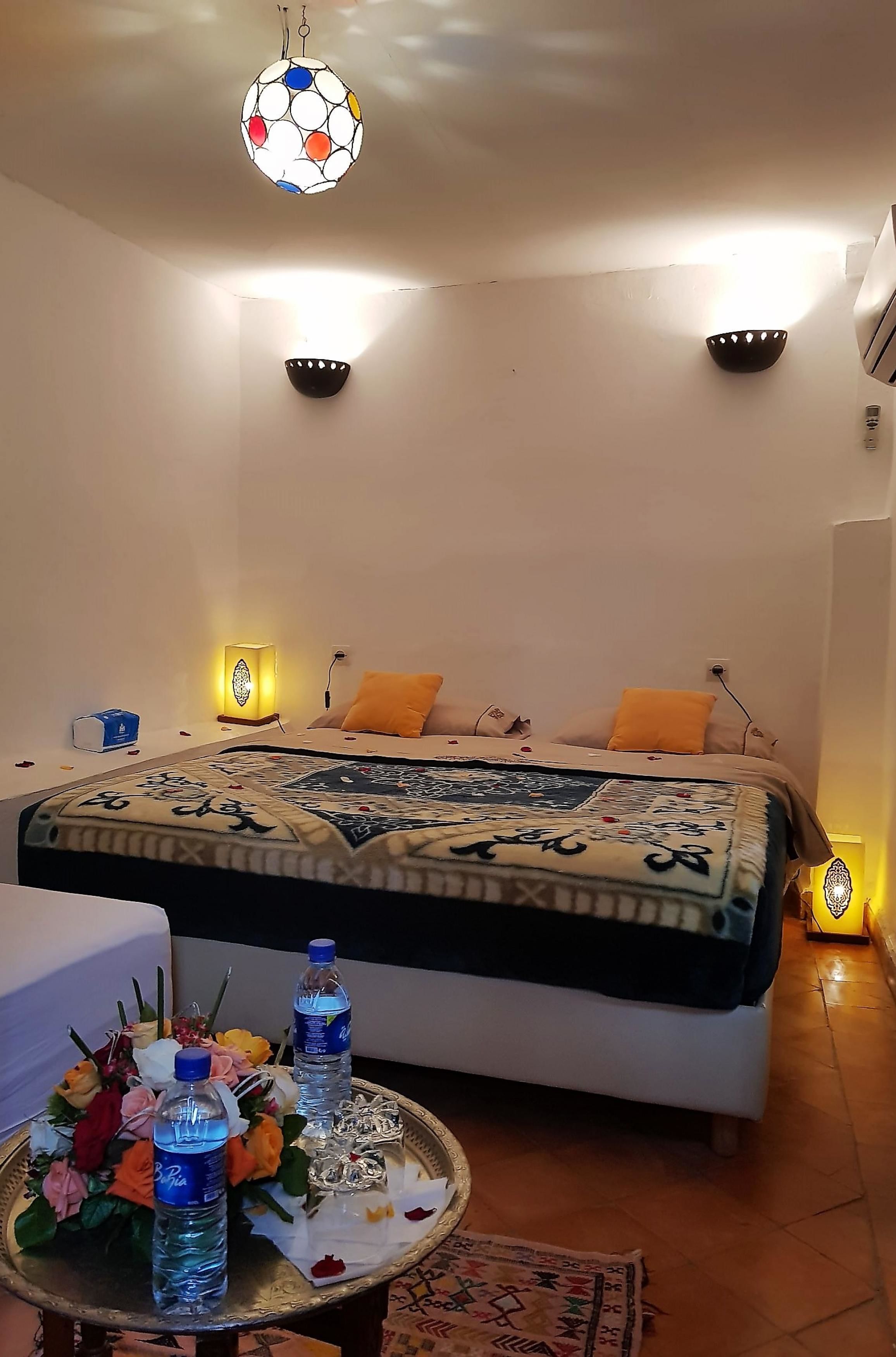 Une chambre à coucher avec un lit, des lampes de chevet, un luminaire décoratif au plafond, une table de chevet avec des bouteilles d’eau et des fleurs, et un tapis à motifs.