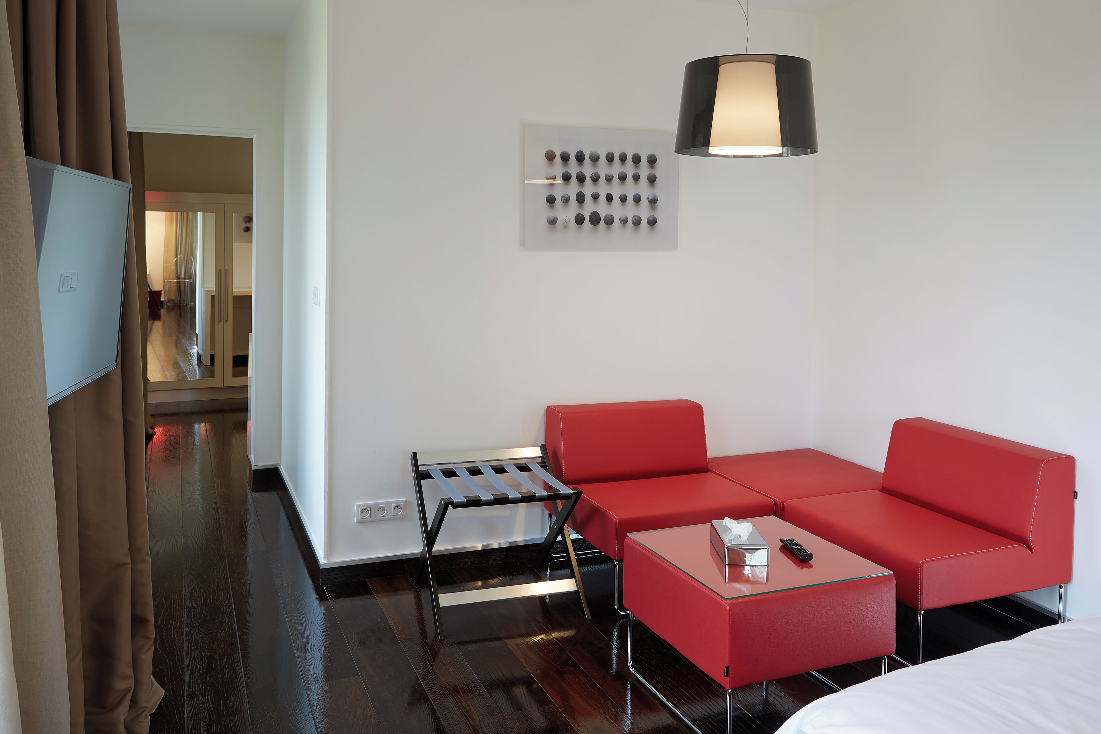 Una habitación de hotel moderna con un sofá rojo, una mesita, un televisor y una lámpara fijada a la pared.