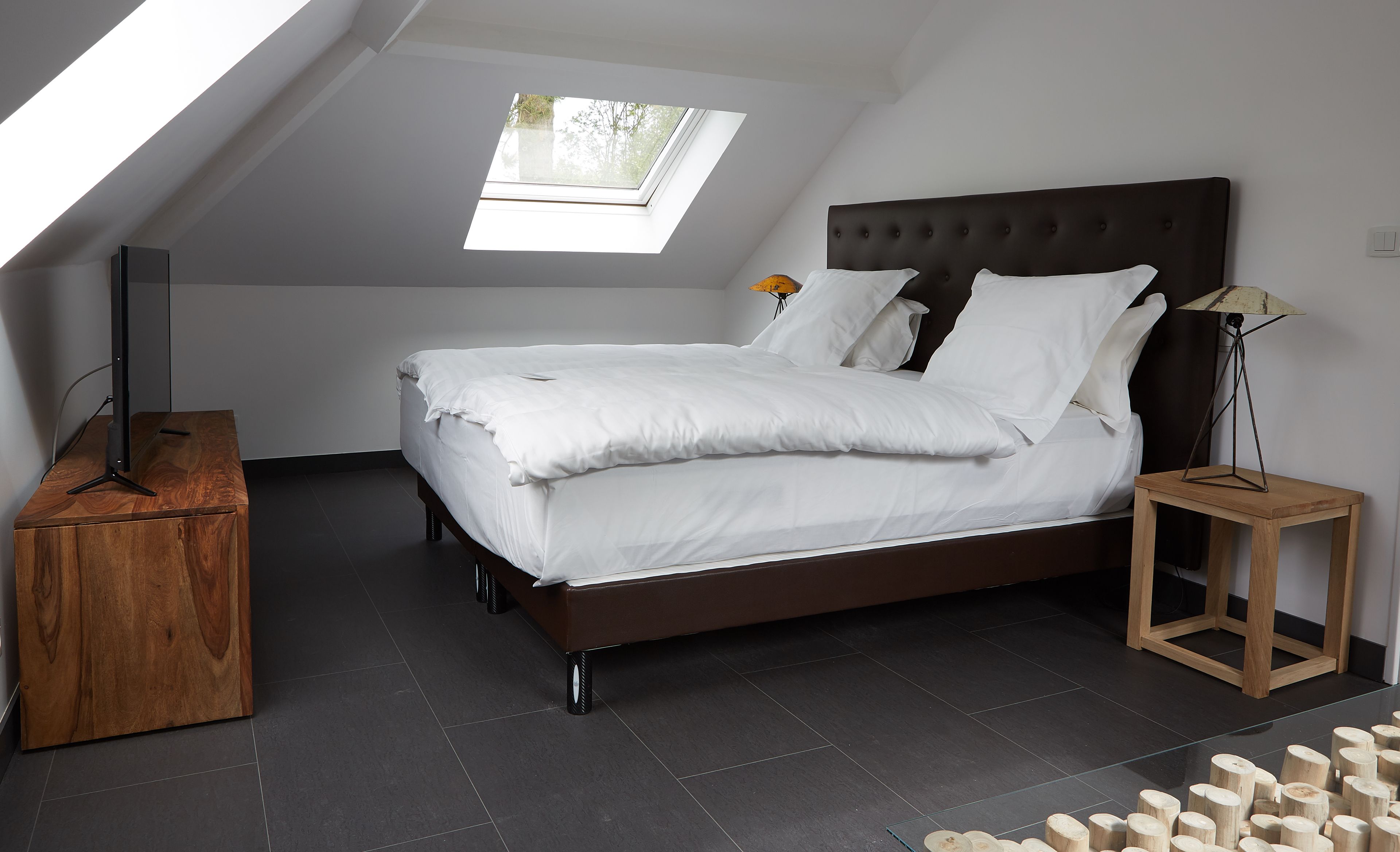 Un dormitorio con techo inclinado, tragaluz, cama con ropa de cama blanca, mesita de noche de madera y lámpara de pie.