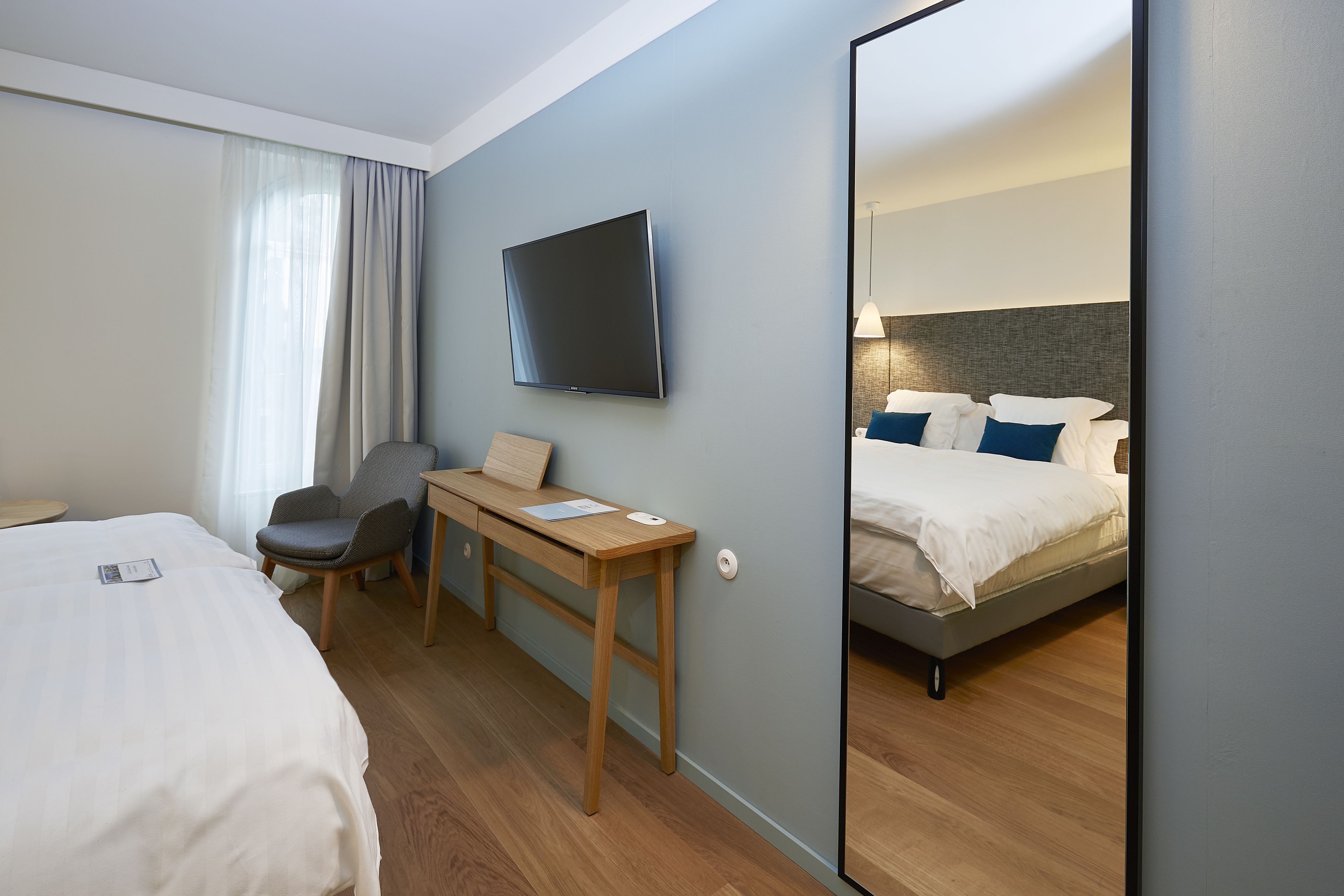 A hotel room with a bed, a desk with a chair, a flat-screen TV, and a large mirror.