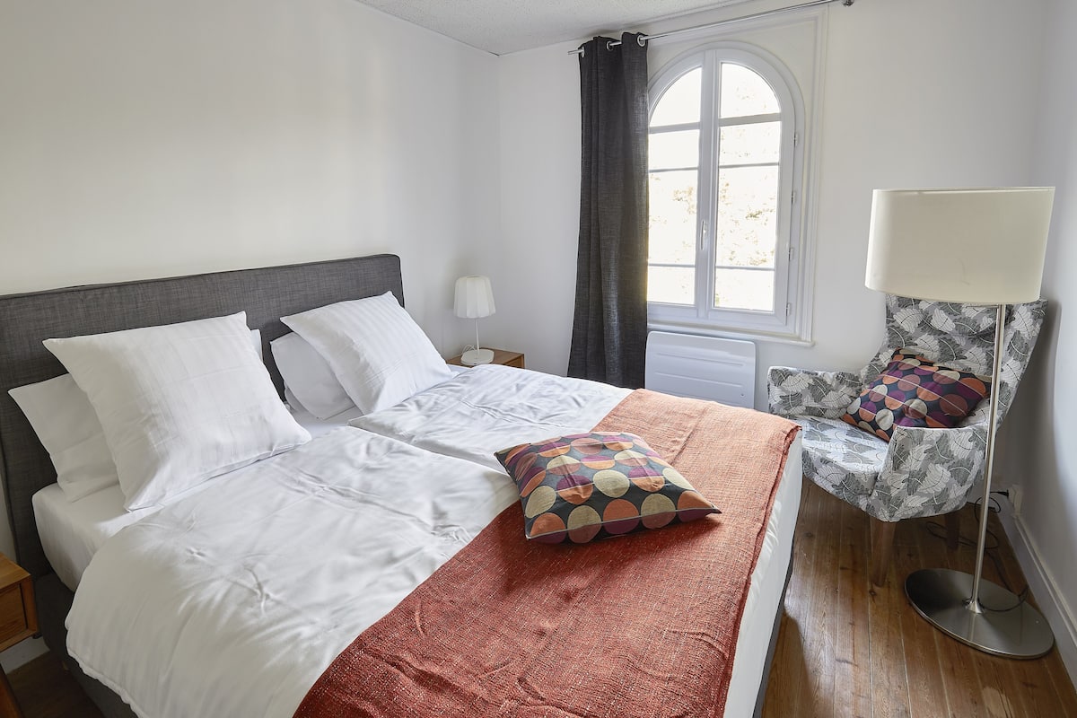 Una cama bien tendida con sábanas blancas y un cojín colorido, una lámpara de noche y un sillón estampado con un cojín.