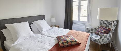 A neatly made bed with white linens and a colorful pillow, a bedside lamp, and a patterned chair with a cushion.