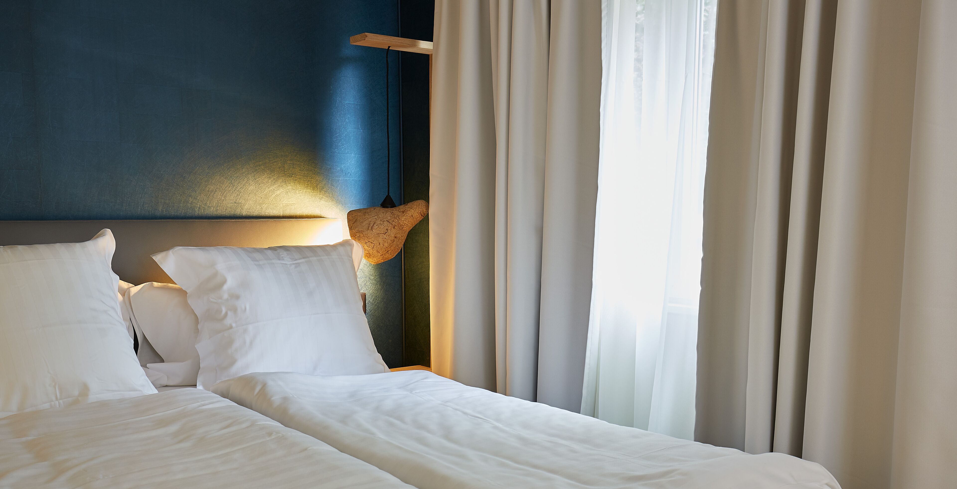 A hotel room with a bed, white bedding, a headboard with a light fixture, and sheer curtains.