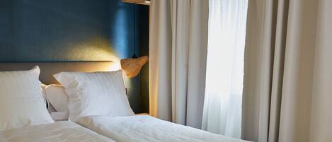 A hotel room with a bed, white bedding, a headboard with a light fixture, and sheer curtains.