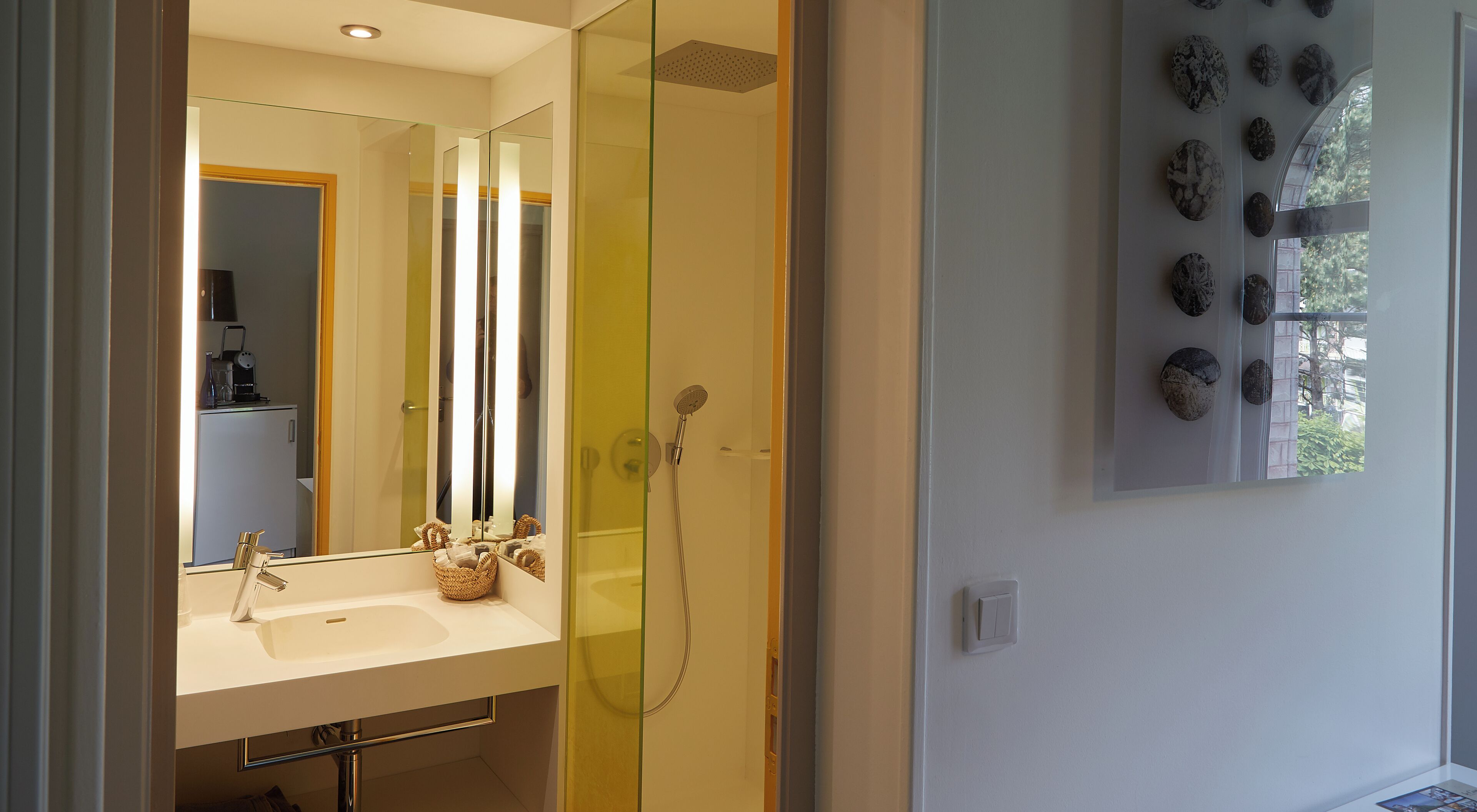 A modern bathroom with a glass shower enclosure, a white sink, and a mirror reflecting a kitchen area.