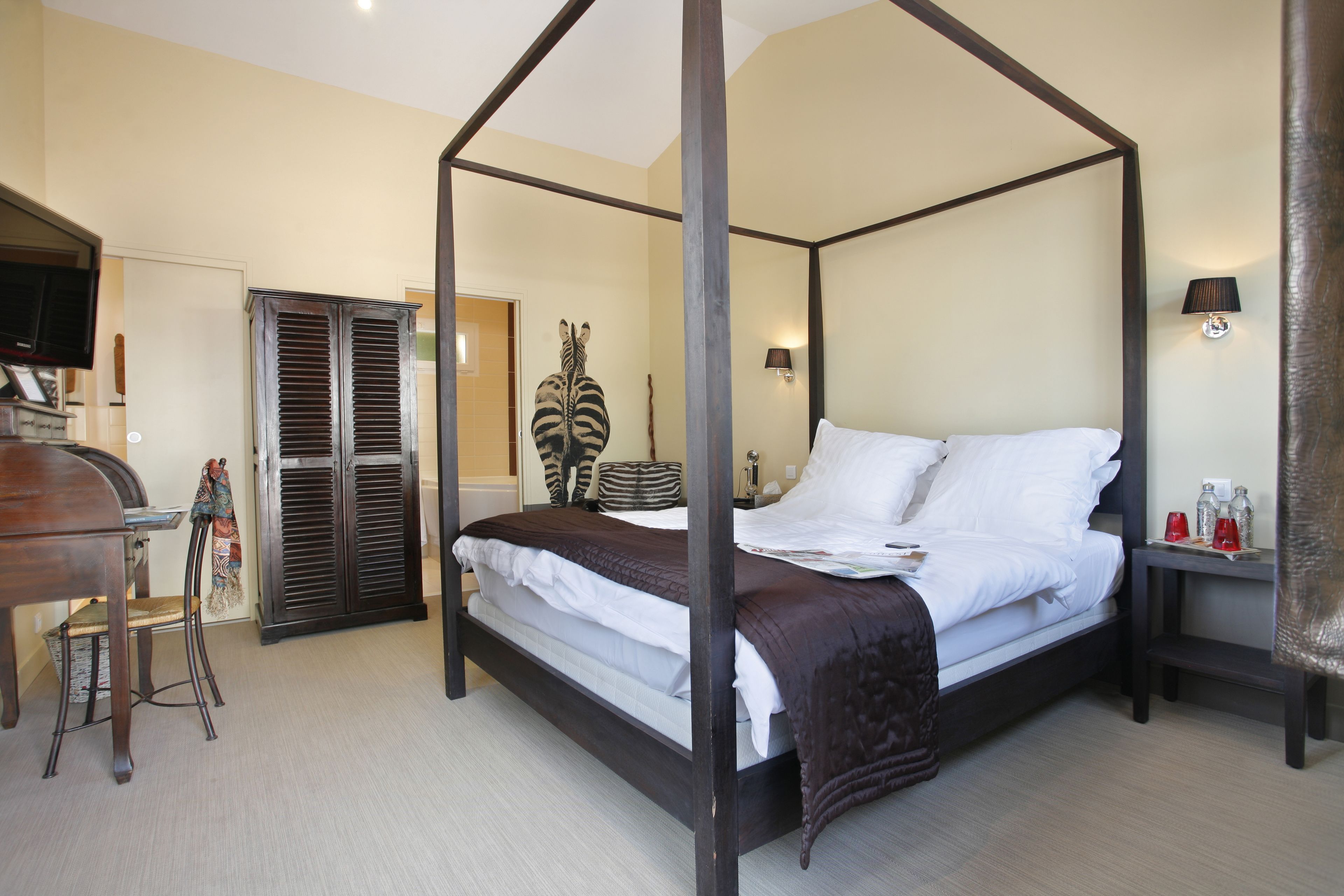 A four-poster bed with a dark frame, white bedding, and a brown throw blanket. There is a wooden desk with a chair, a television, and a decorative item on the wall.