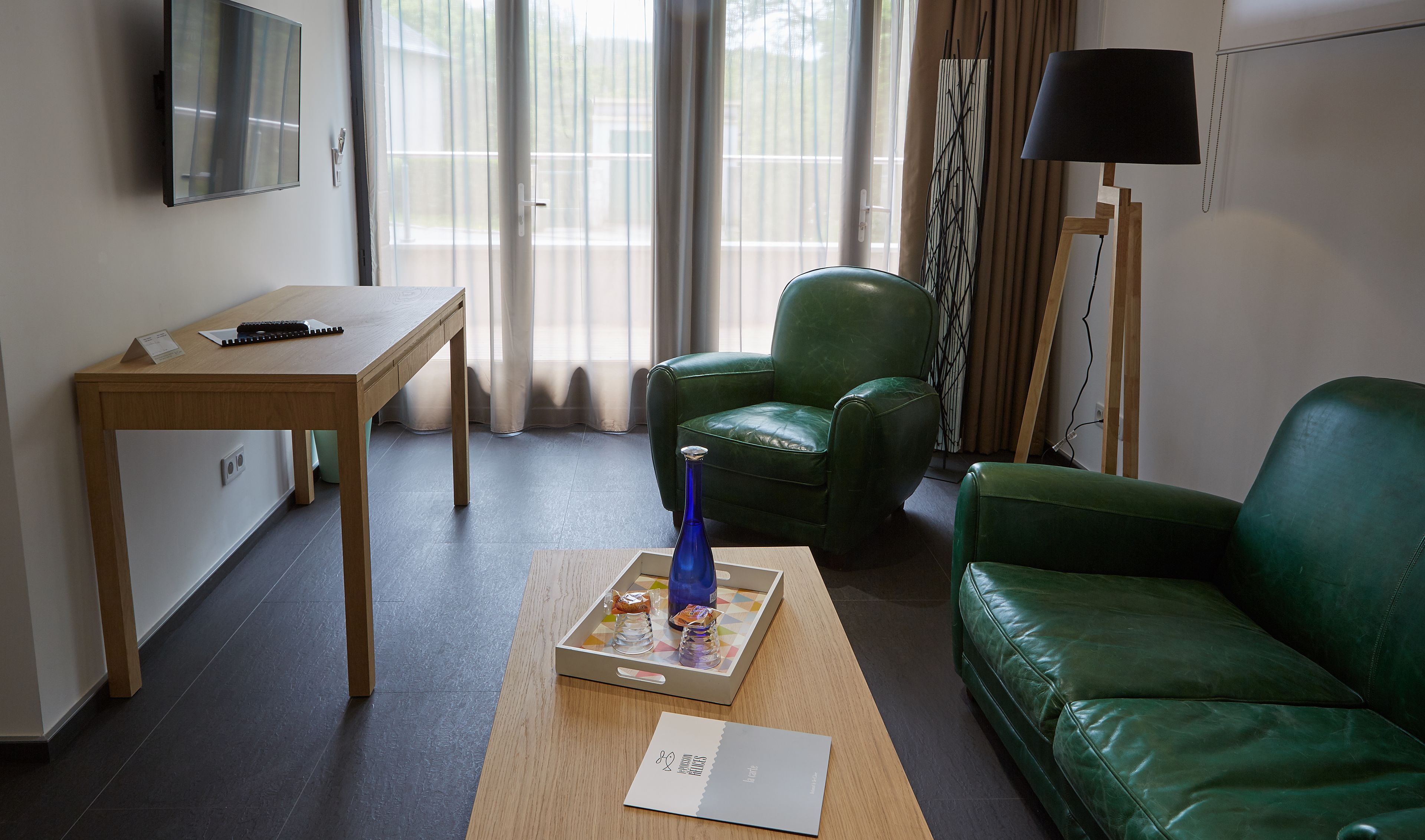 Ein modernes Wohnzimmer mit einem Holztisch, einem grünen Ledersofa und einem grünen Sessel. Auf dem Boden stehen eine blaue Vase und ein Tablett mit Gläsern.