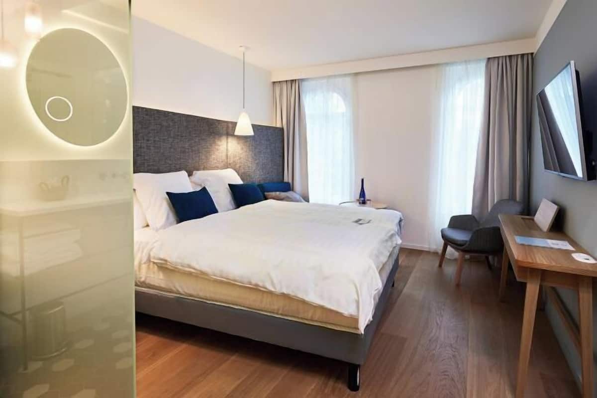 Una habitación de hotel moderna con una cama grande, un escritorio, una silla, un televisor y un espejo montado en la pared.