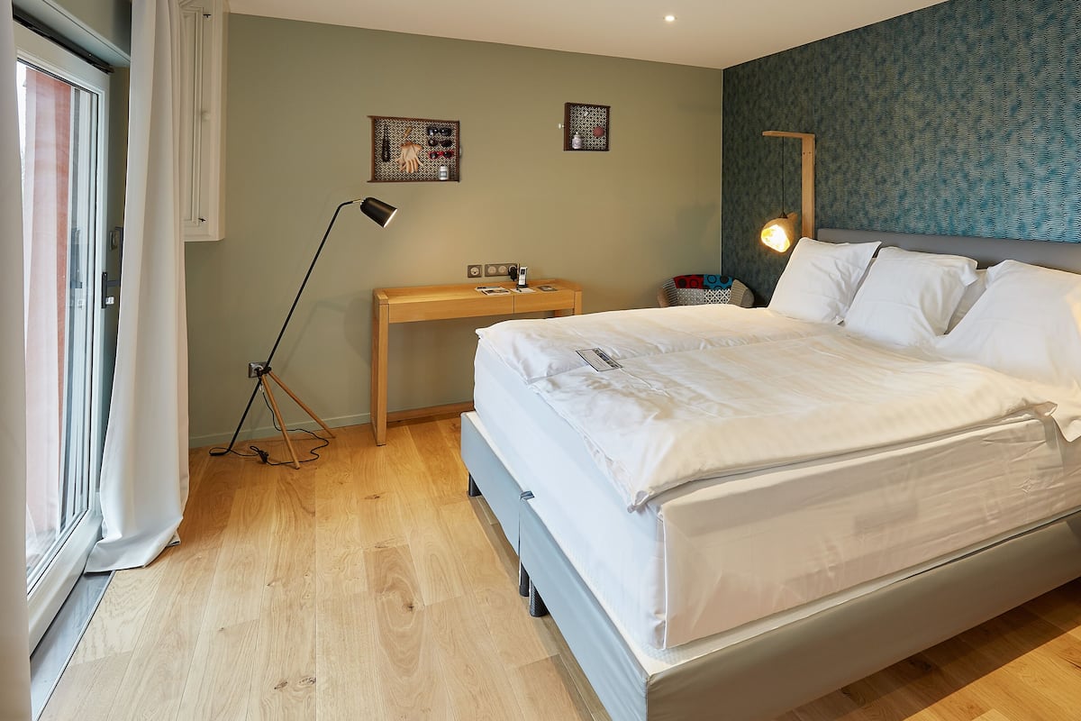 Una cama bien tendida en una habitación con suelo de madera, un escritorio y un espejo en la pared.