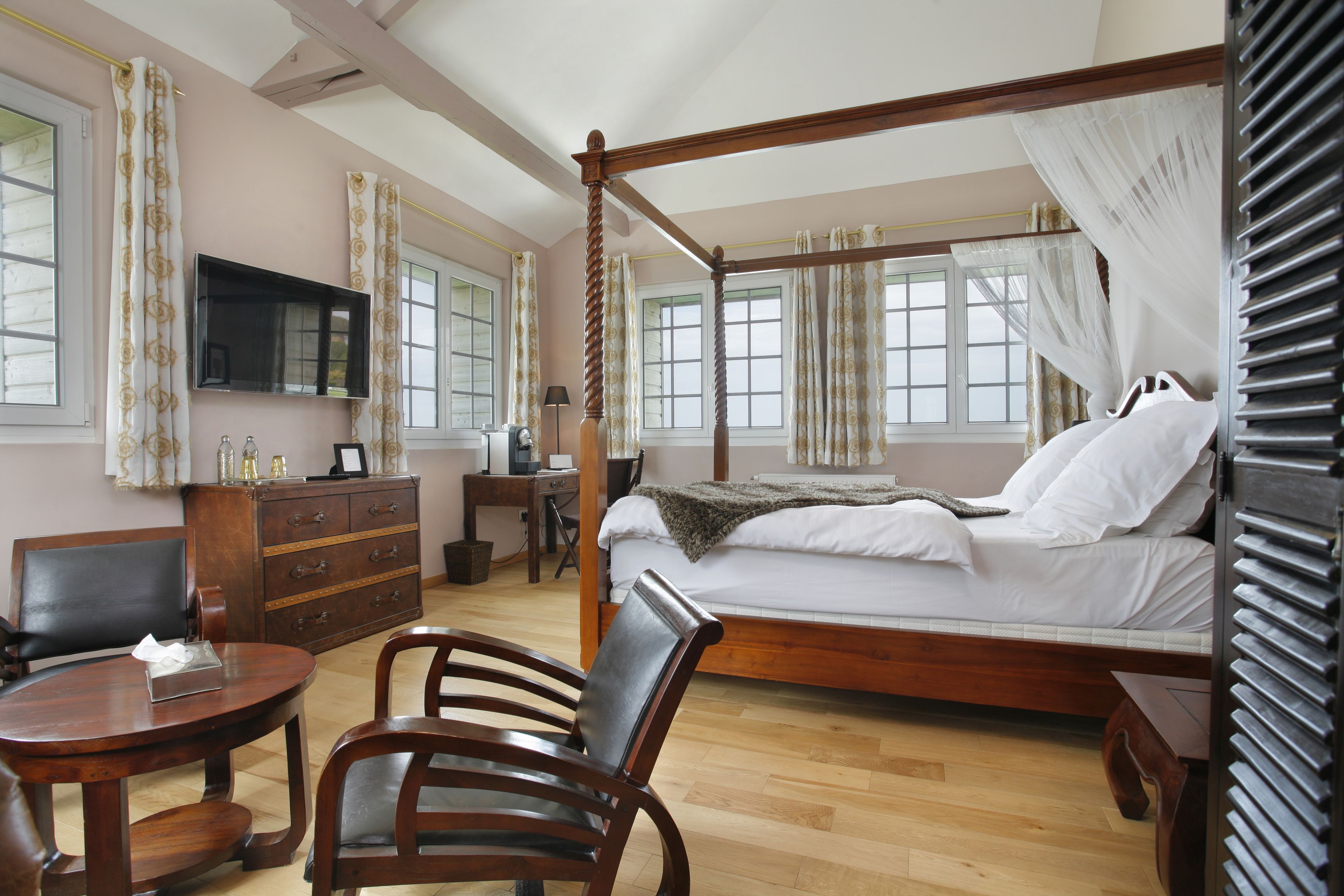 A four-poster bed with a canopy, a television mounted on the wall, a desk with a chair, and a small round table with a chair.