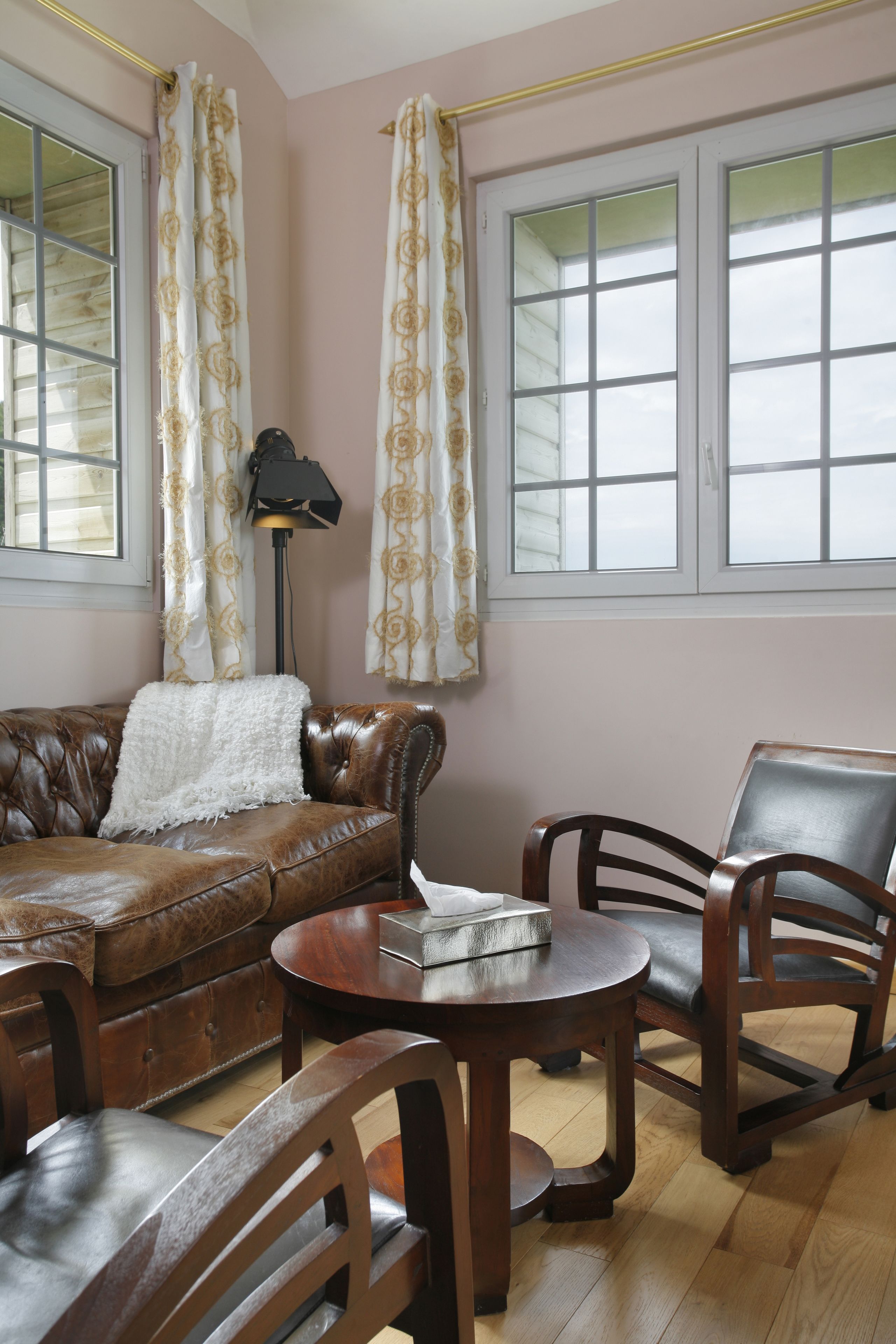 Une pièce avec un canapé en cuir marron, une table basse en bois et deux chaises en bois. Il y a une fenêtre avec des rideaux blancs et une lampe sur un pied.