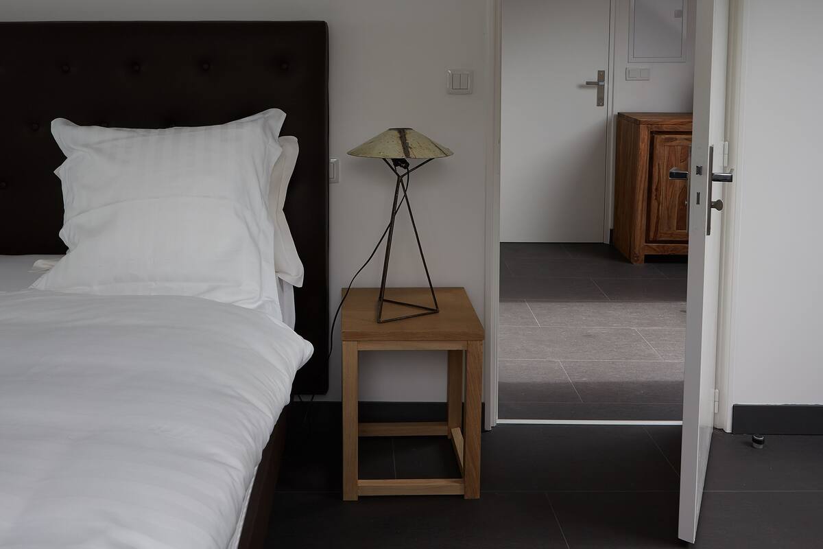 Un dormitorio con una cama, una mesita de noche con lámpara, y una puerta que da a otra habitación.