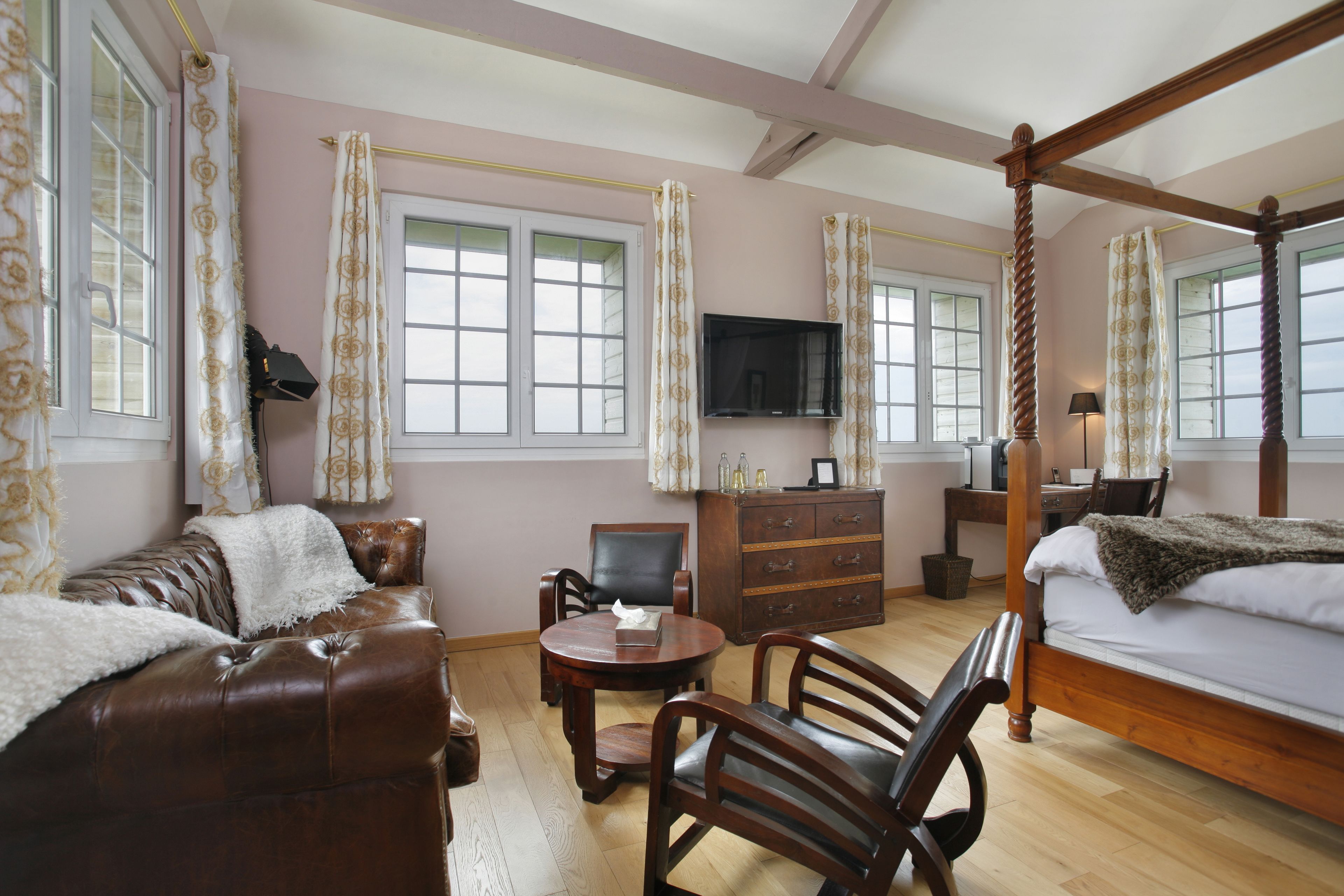 A bedroom with a four-poster bed, a wooden dresser, a television, and a seating area with a sofa and chairs.