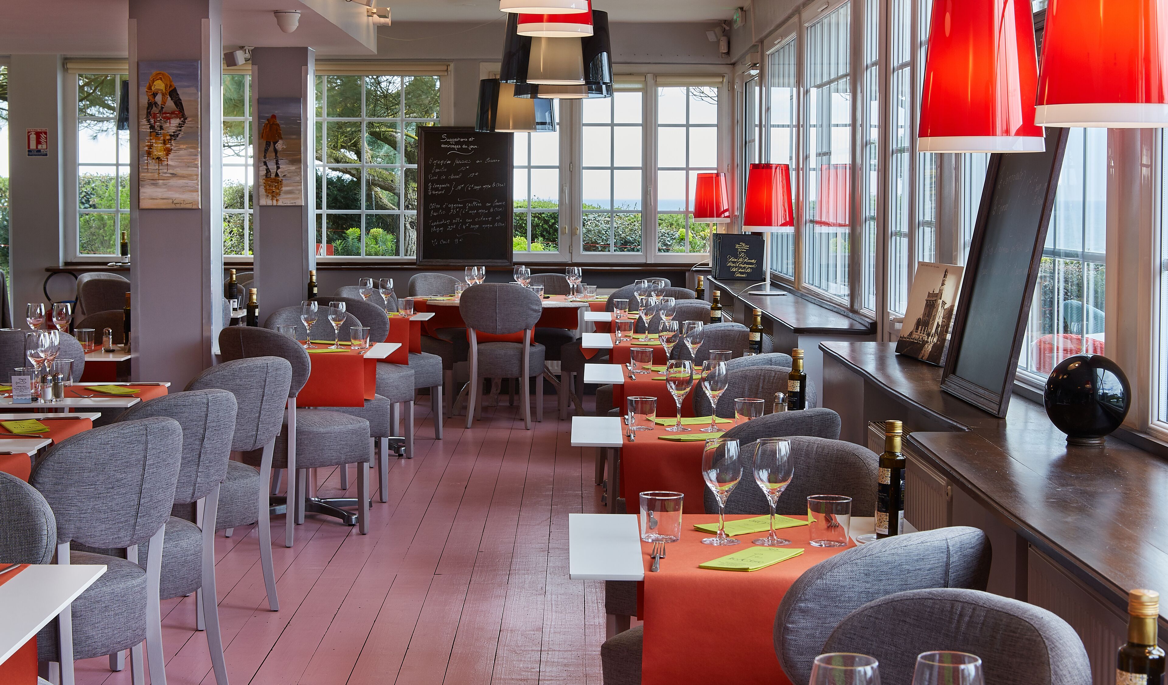 A restaurant with tables set for dining, large windows, and red pendant lights.