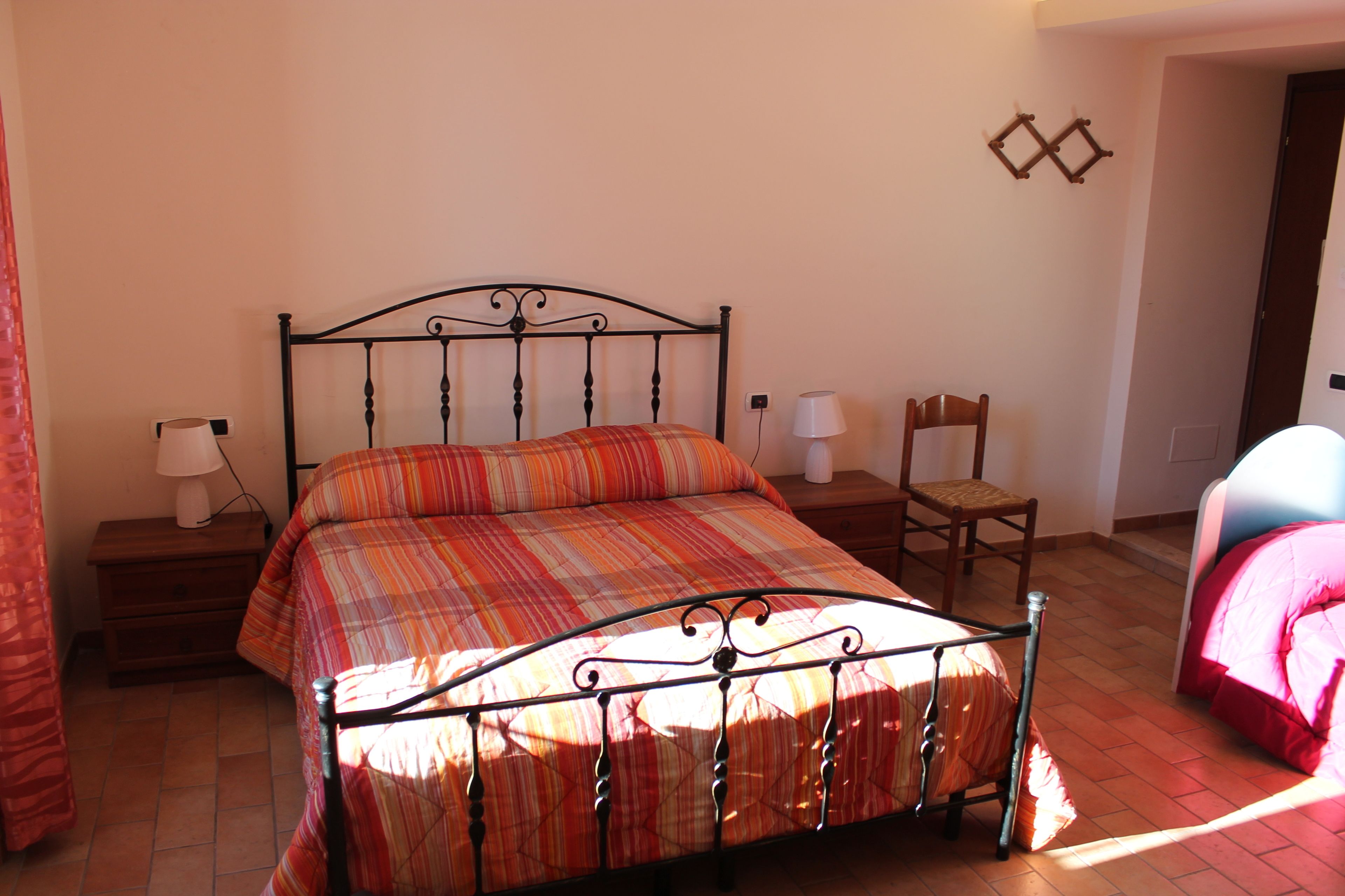 Un dormitorio con estructura de cama metálica, colcha escocesa, mesita de noche de madera con lámpara, una silla y una puerta que da a otra habitación.
