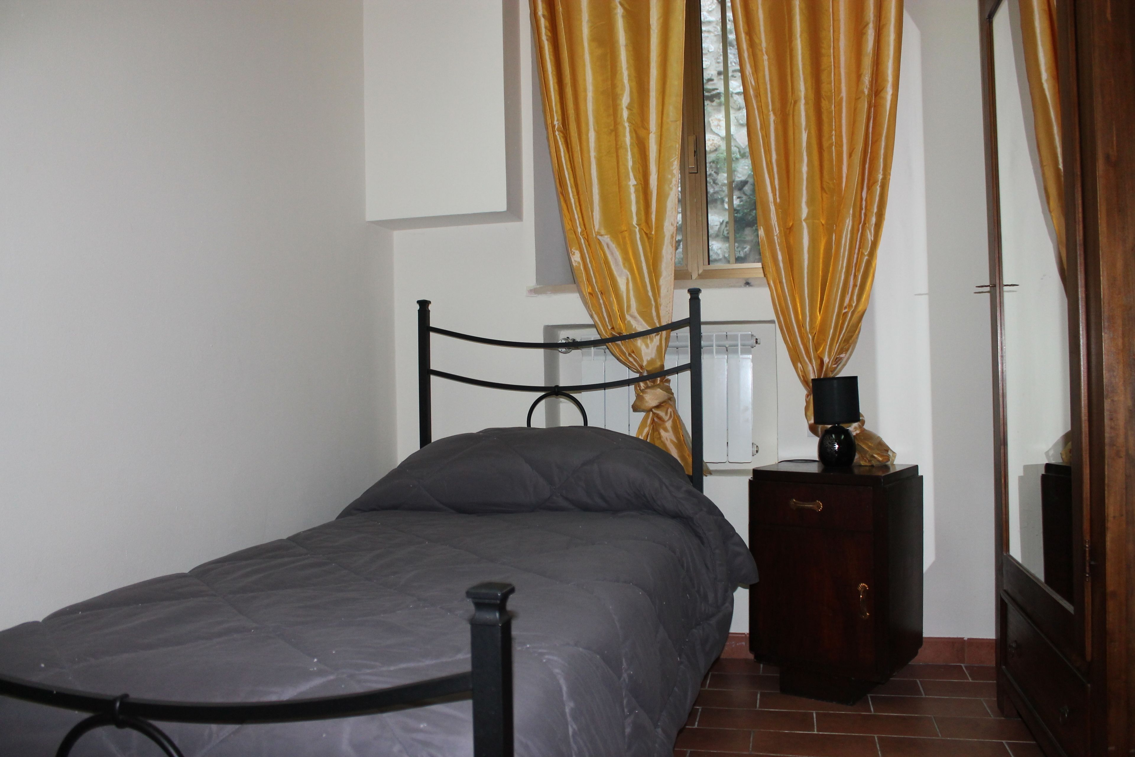 A single bed with a metal frame, a bedside table with a lamp, and a window with yellow curtains.
