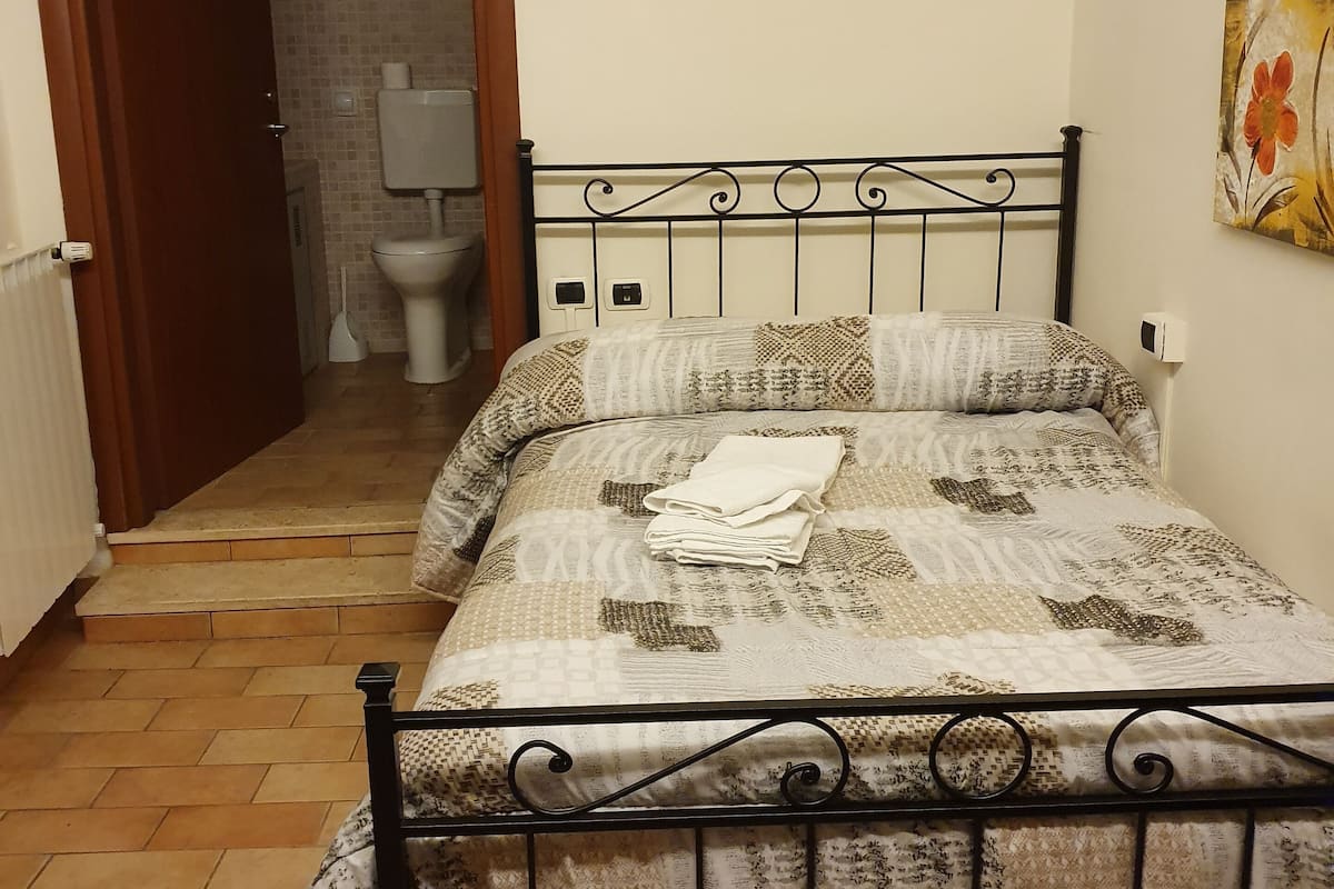 A bedroom with a bed, a bathroom visible through an open door, and a painting on the wall.