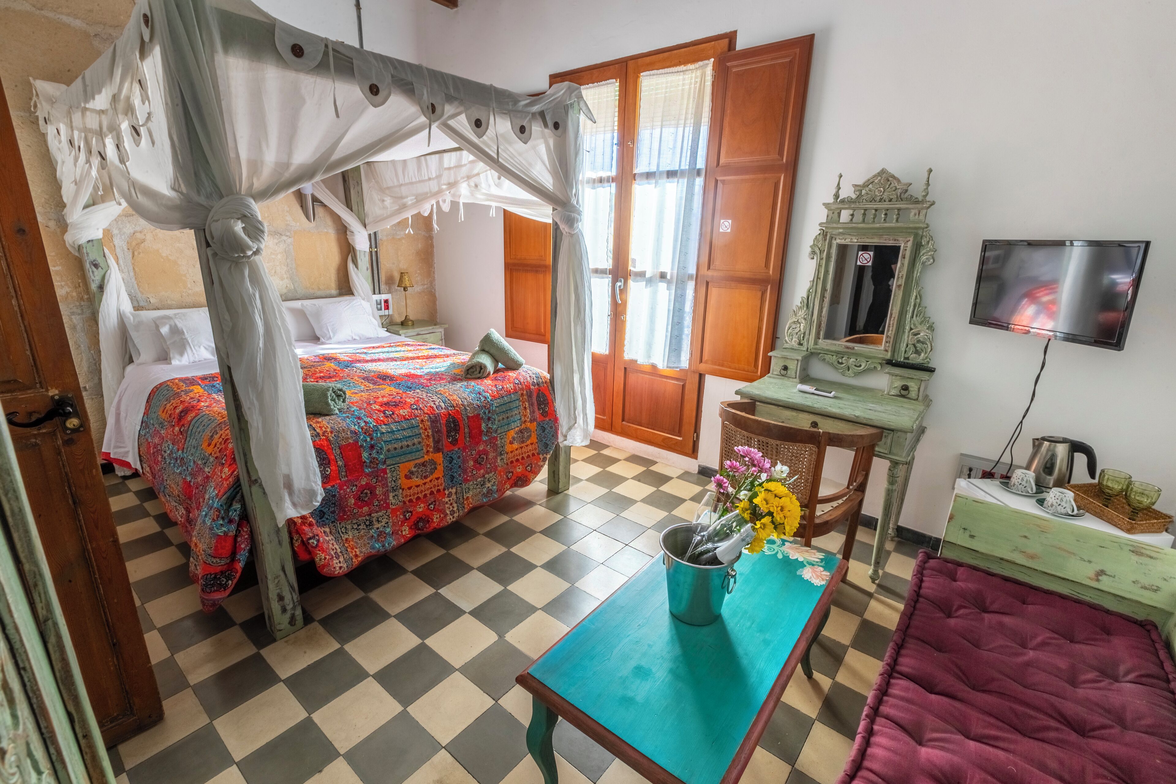 Una habitación con una cama con dosel, suelo a cuadros, una mesa con un florero y un televisor en la pared.