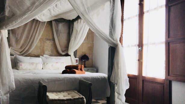 A bed with a canopy, a nightstand, a lamp, and a chair in a room with a wooden door and a window. - Ibiza Boutique Guest House (Ibiza Town)