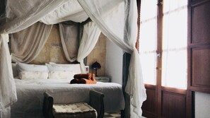 A bed with a canopy, a nightstand, a lamp, and a chair in a room with a wooden door and a window. - Ibiza Boutique Guest House (Ibiza Town)