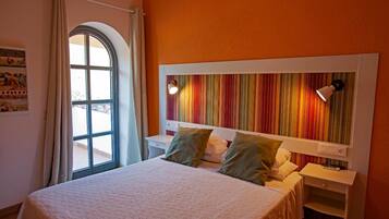 Una habitación de hotel con una cama, una mesita de noche, un ventanal con cortinas y un cabecero a rayas de colores.