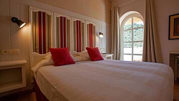 Una habitación de hotel con una cama grande, dos almohadas rojas, una mesita de noche, una lámpara, un ventanal con cortinas y una pared con rayas verticales.