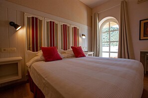 Una habitación de hotel con una cama grande, dos almohadas rojas, una mesita de noche, una lámpara, un ventanal con cortinas y una pared con rayas verticales.