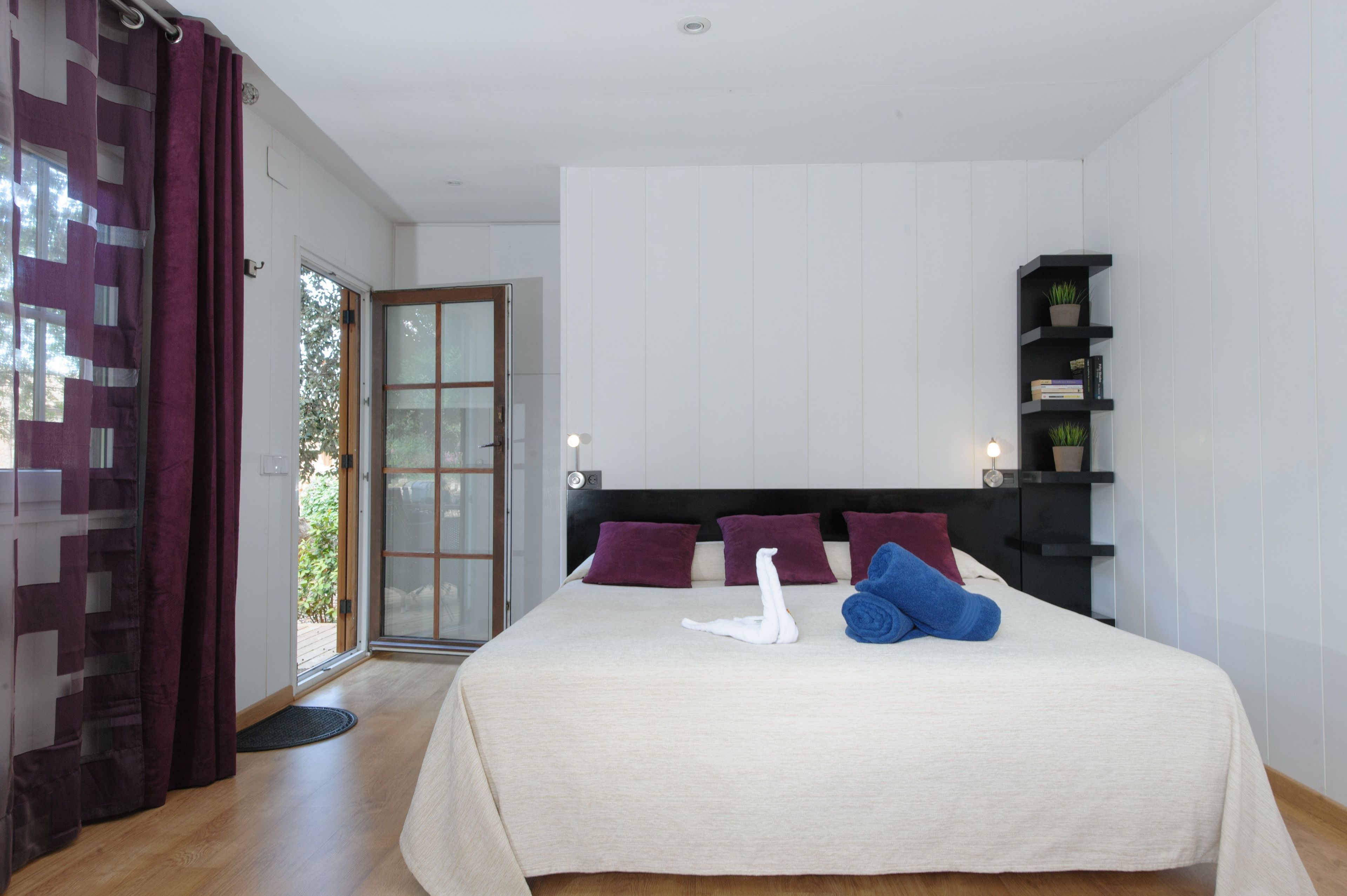 Une chambre à coucher avec un lit, des oreillers, une étagère noire ornée de plantes et une porte-fenêtre coulissante donnant sur un espace extérieur.