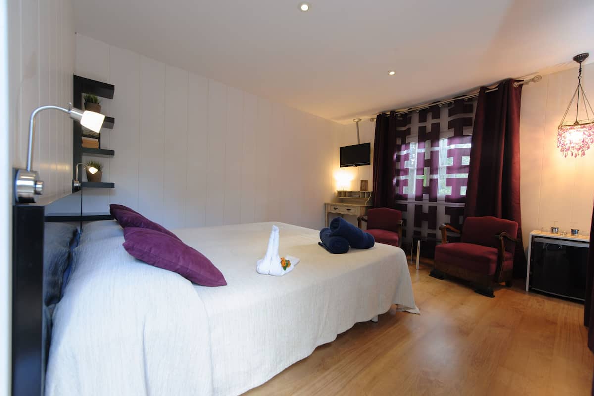 A neatly made bed with pillows, a bedside lamp, a flat-screen TV, a small table with a lamp, and a chair.