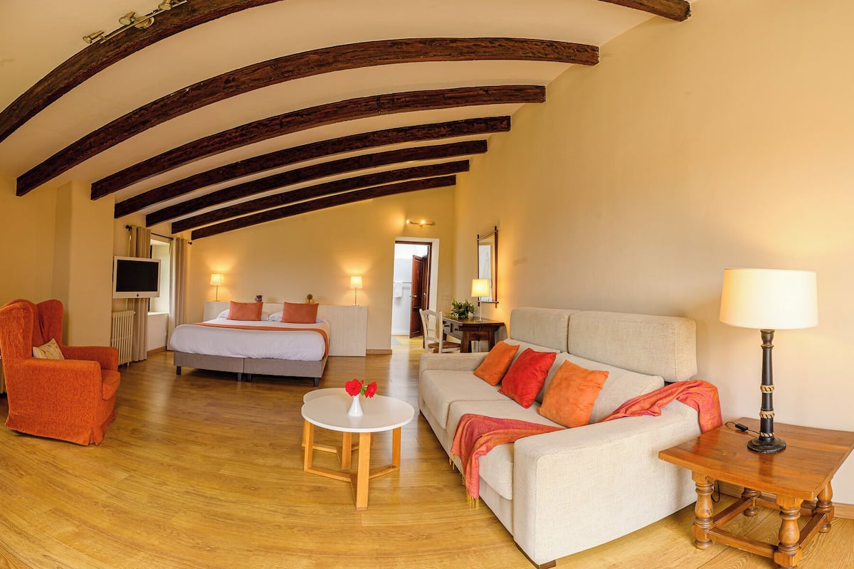 A spacious room with wooden floors, a bed, a sofa, a chair, and a small table with a lamp.